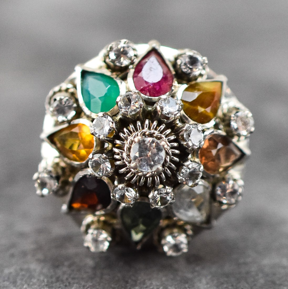 10K GOLD MULTI-GEMSTONE 'THAI PRINCESS' RING W/ RUBY, EMERALD, TOPAZ, TIGER'S EYE: A 10K white gold 'Thai Princess' ring with a crown of many precious and semi-precious gemstones including ruby, emerald, topaz, quartz, tiger's eye, and more, circa 1960s-70s. The ring has a weight