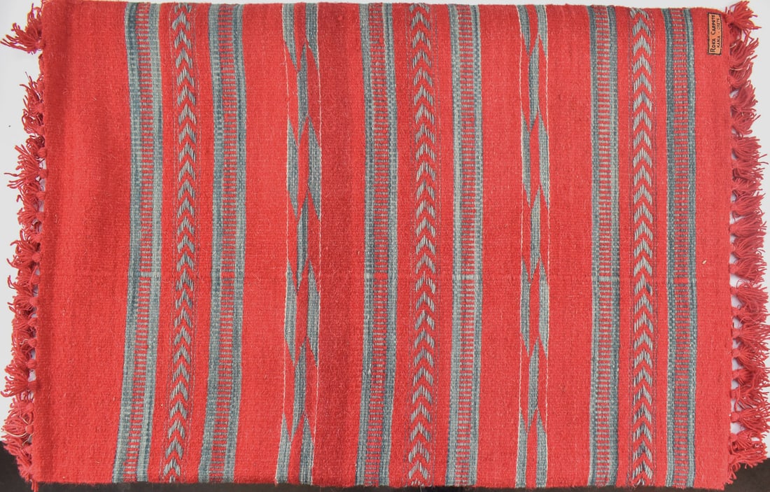 WOOL RUG ON WOOD FRAME, CRETE: A wool rug with a striped pattern and deep red color originating from Roka Carpets of Hania, Crete. The maker's tag is displayed on the front of the rug. The rug on the wood frame measures 28" H x