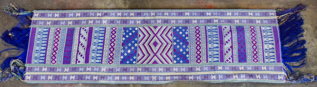 SILK RUNNER RUG ON A WOOD FRAME, BHUTAN: A silk runner rug on a wood with pink, blue, and purple colors and geometric designs, likely originating from Bhutan. The rug on the frame measures 24" H x 81" W x 1" D.