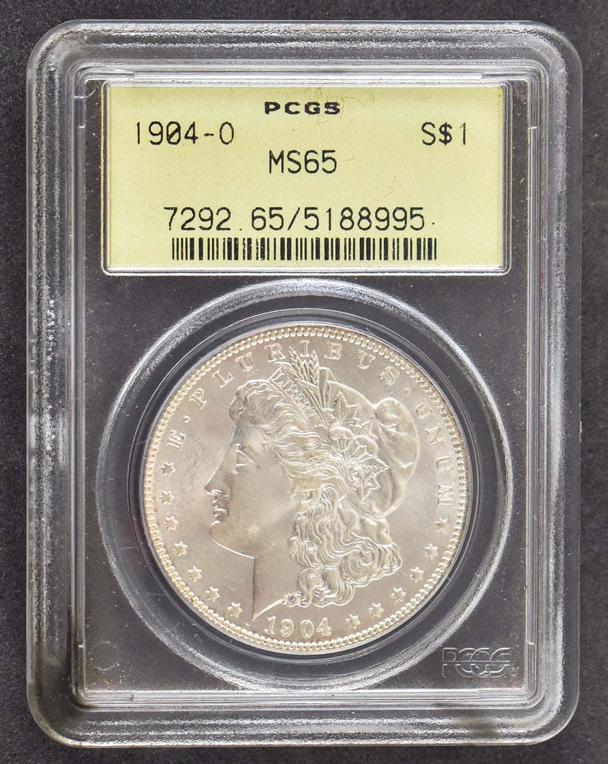 1904-O MORGAN SILVER DOLLAR PCGS MS65: A 1904-O Morgan silver dollar graded by PCGS a Mint State 65 grade. Held in a hard plastic PCGS slab measuring 3.25" H x 2.5" W x 0.35" D. The coin is struck in 90% silver and has a weight of approxim