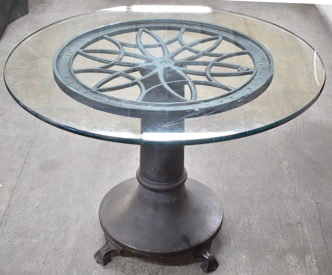 THE SWIFT MILL LANE BROTHERS CAST IRON COFFEE MILL WHEEL REPURPOSED TABLE: The Swift Mill Lane Brothers cast iron coffee mill wheel repurposed into a glass top table with a cast iron base, circa late 19th century. The item measures 30" Hx 42" W x 42" D.