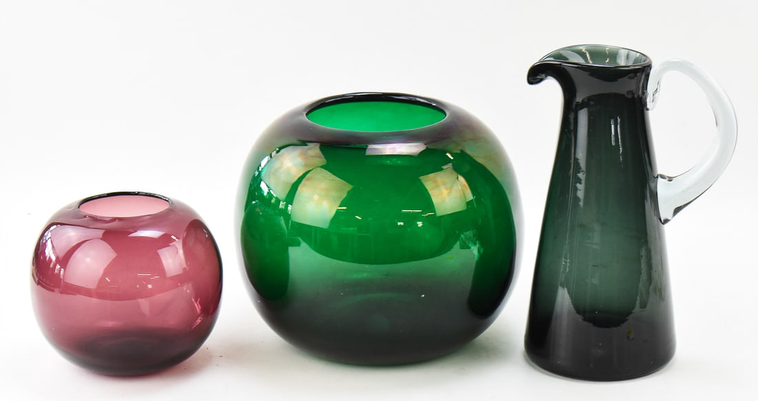 TRIO OF MID-CENTURY EMERALD GREEN & AMETHYST BLENKO ART GLASS: A trio of Mid-century hand-blown Blenko art glass which includes a round amethyst vase, a round emerald green vase, and a emerald green with clear handle pitcher measuring 10" H x 5" W x 5" D.