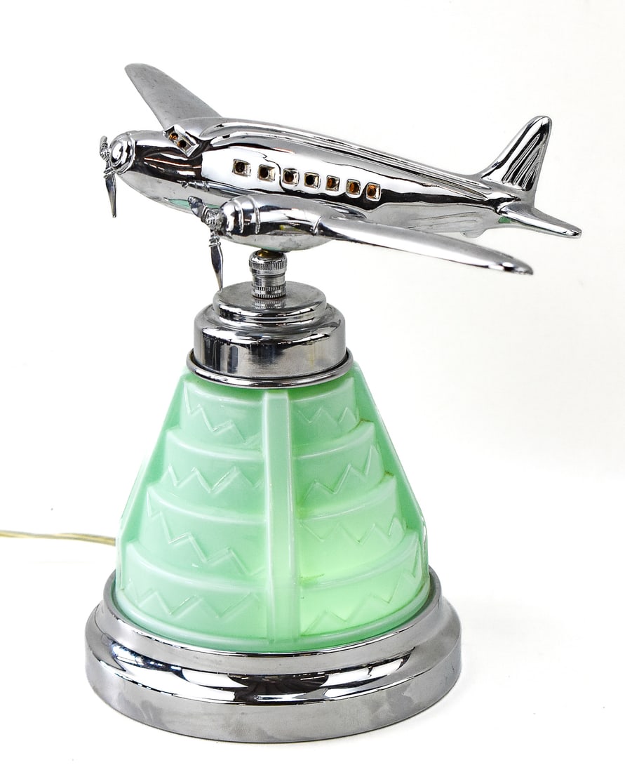 DERECO CLASSICS ART DECO STYLE CHROME DC-2 AIRPLANE ACCENT LAMP: An Art Deco style chrome DC-2 airplane accent lamp by Dereco Classics. The lamp has a large pivoting plane on top with spinning propellors, frosted green stepped-up base; and a three-way switch which 