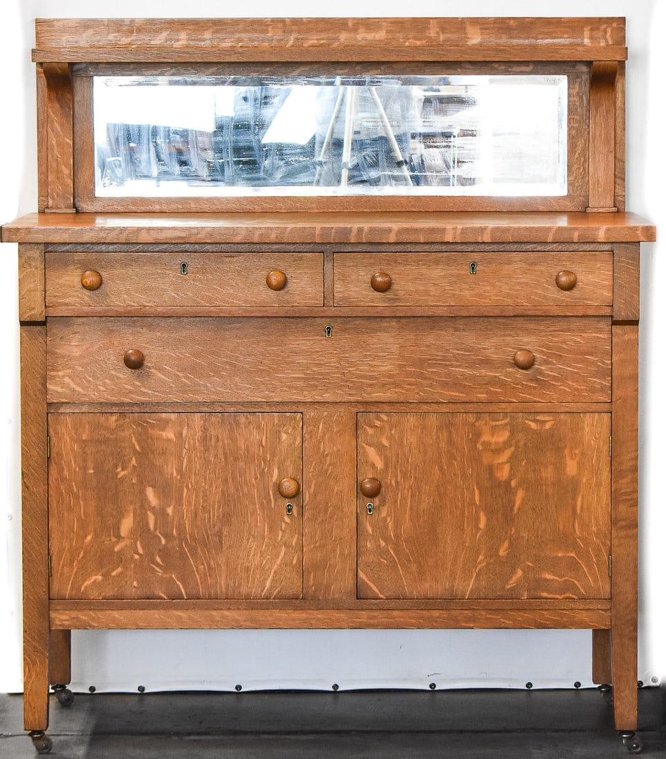 ARTS AND CRAFTS OAK SIDEBOARD: An Arts and Crafts oak mirrored sideboard with three drawers and is on casters, circa early 20th century. The item measures 55.5" H x 48" W x 22.5" D.