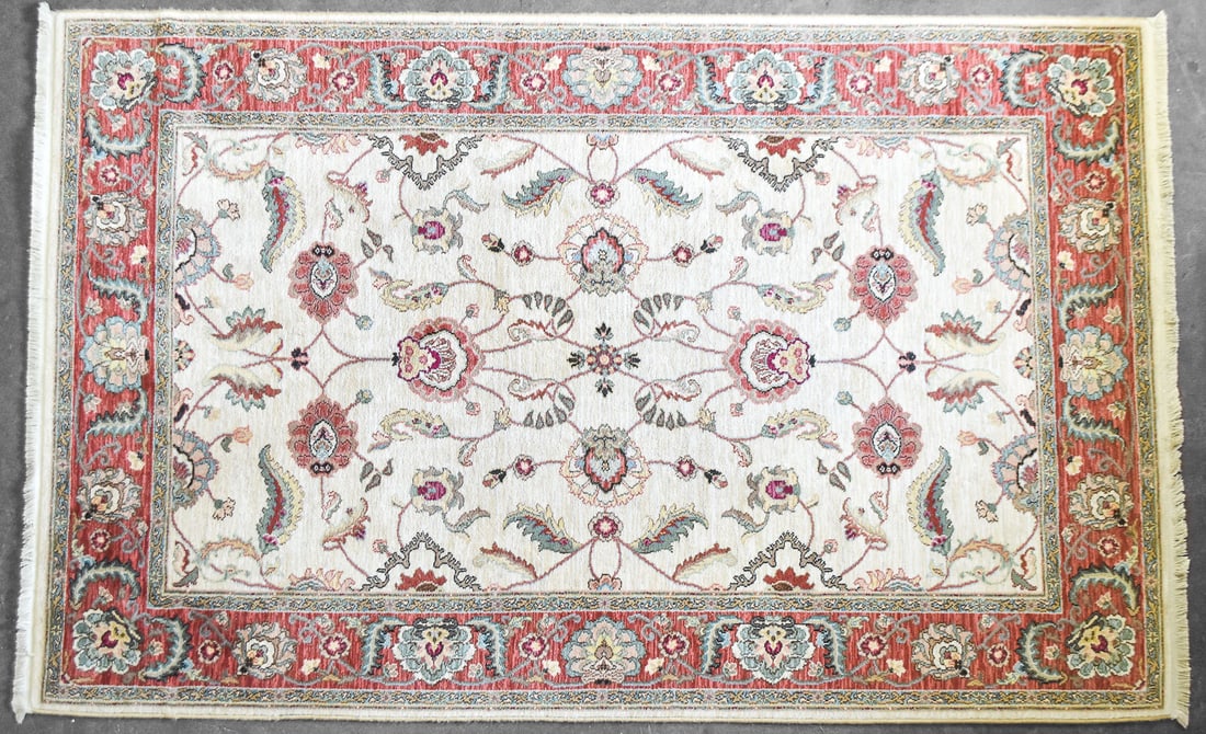 KARASTAN ASHARA "AGRA IVORY" PATTERN WOOL RUG: A Karastan Ashara 100% wool rug in the "Agra Ivory" pattern with a traditional floral design in the field and a thick red border. The rug measures 112" H x 70" W x 0.5" D.