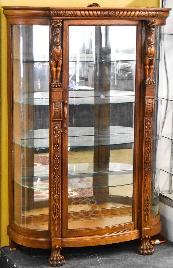 HORNER STYLE CURVED GLASS DISPLAY CABINET: R.J. Horner style curved glass display cabinet featuring heavy carved lions on the pilasters and lion paw feet. Cabinet is made of quarter sawn tiger oak with bowed glass front and glass adjustable