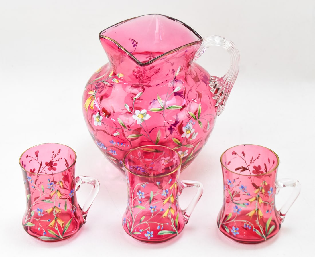 VICTORIAN CRANBERRY GLASS ENAMELED JUICE SET: A Victorian cranberry glass pitcher with an enameled flower motif and three matching handled cups. Pitcher measures 7" H x 6"W x 6"D