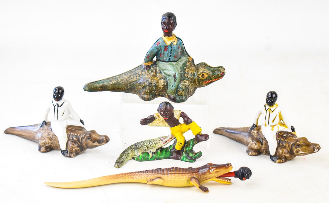 BLACK AMERICANA ALLIGATOR FIGURINES: CAST IRON, PORCELAIN, CELLULOID: An assortment of five alligator design Black Americana figurines including a celluloid alligator pencil holder circa 1940s-1950s, two porcelain figures riding on an alligator made circa mid-20th