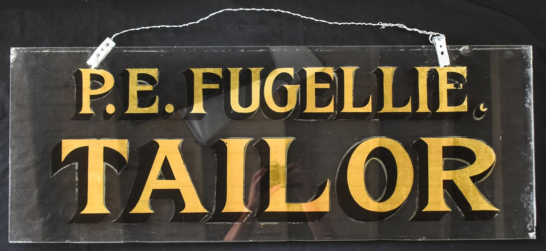 P.E. FUGELLIE REVERSE-PAINTED GLASS TAILOR ADVERTISEMENT SIGN: A P.E. Fugellie (Lander, WY) reverse-painted glass tailor advertisement sign with gold-leaf lettering, from the early 1900s. The sign measures approx. 13.5" H x 36" W x 0.5" D.