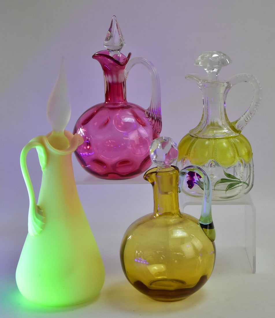ASSORTMENT OF FOUR GLASS CRUETS: An assortment of four glass cruets with stoppers including a Burmese uranium glass cruet, a cranberry glass coin-dot design cruet with a twisted clear glass handle, an amber glass cruet with clear