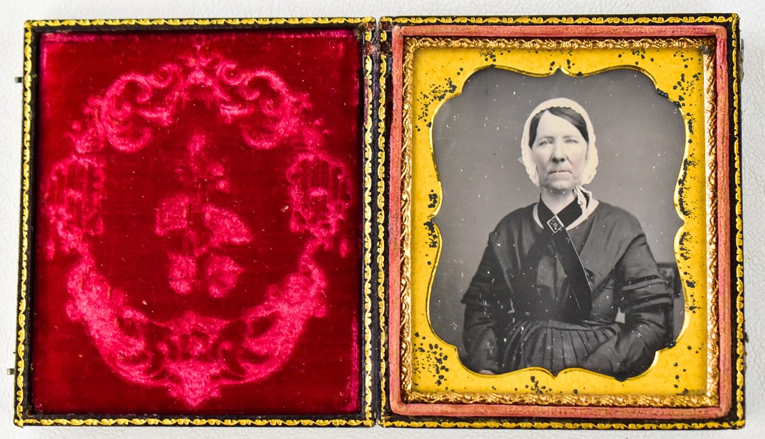 DAGUERREOTYPE OF A WOMAN IN BLACK: A daguerreotype of an older woman with a detailed neck piece in a 1/6 plate leather case, circa 1854. The case measures 4" H x 3" W x 0.5" D when closed.