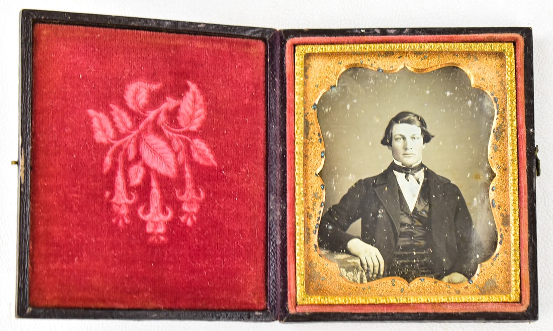 DAGUERREOTYPE OF SUITED YOUNG MAN: A daguerreotype of a suited young man in a leather 1/6th plate case featuring drooping fuchsias, circa 1850s. The case measures 4" H x 3" W x 0.5" D.