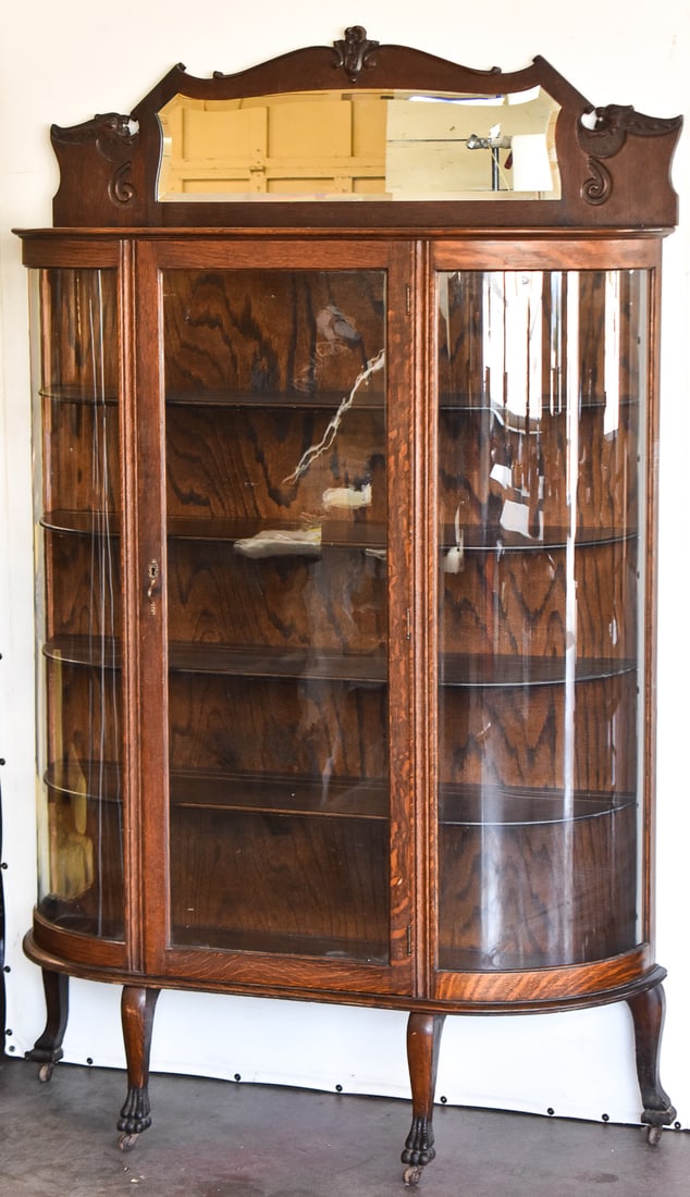 AMERICAN EMPIRE BOW FRONT CURIO CABINET: This curio cabinet is made of quarter sawn tiger oak circa 1900s. It has bend glass sides, claw foot feet with wheels, and American Empire scroll work detail around the top mirror. Cabinet measures