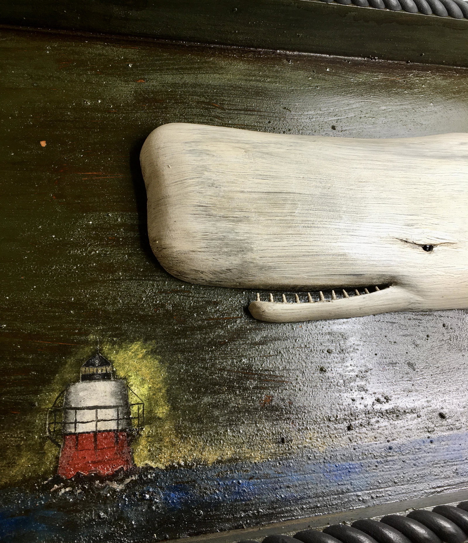 Carved White Whale with Hand Painted Frame: A 14 3/8 by 41 3/4 by 3 1/4 wooden rope edged frame displaying a 25 inch carved white sperm whale plaque and a lighthouse painting by Roger and Brian Mitchell. Signed on the back.In original paint wit