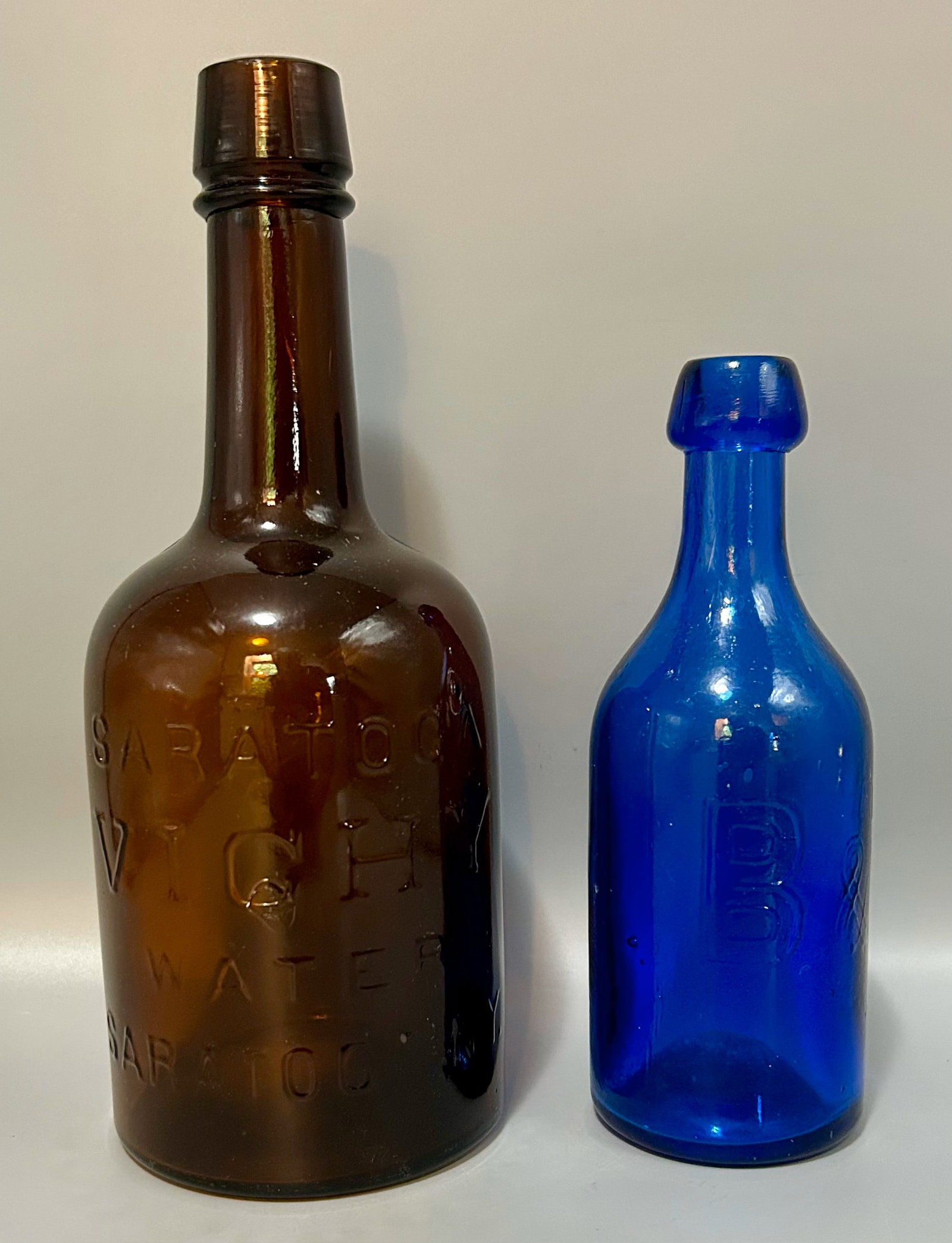 A Saratoga Vichy Water and a B & T Soda Bottle: A 10 inch brown bottle by Saratoga Vichy Water, Saratoga, NY and a 7 1/4 inch blue soda bottle by "B & T".As found- with expected wear for its age,