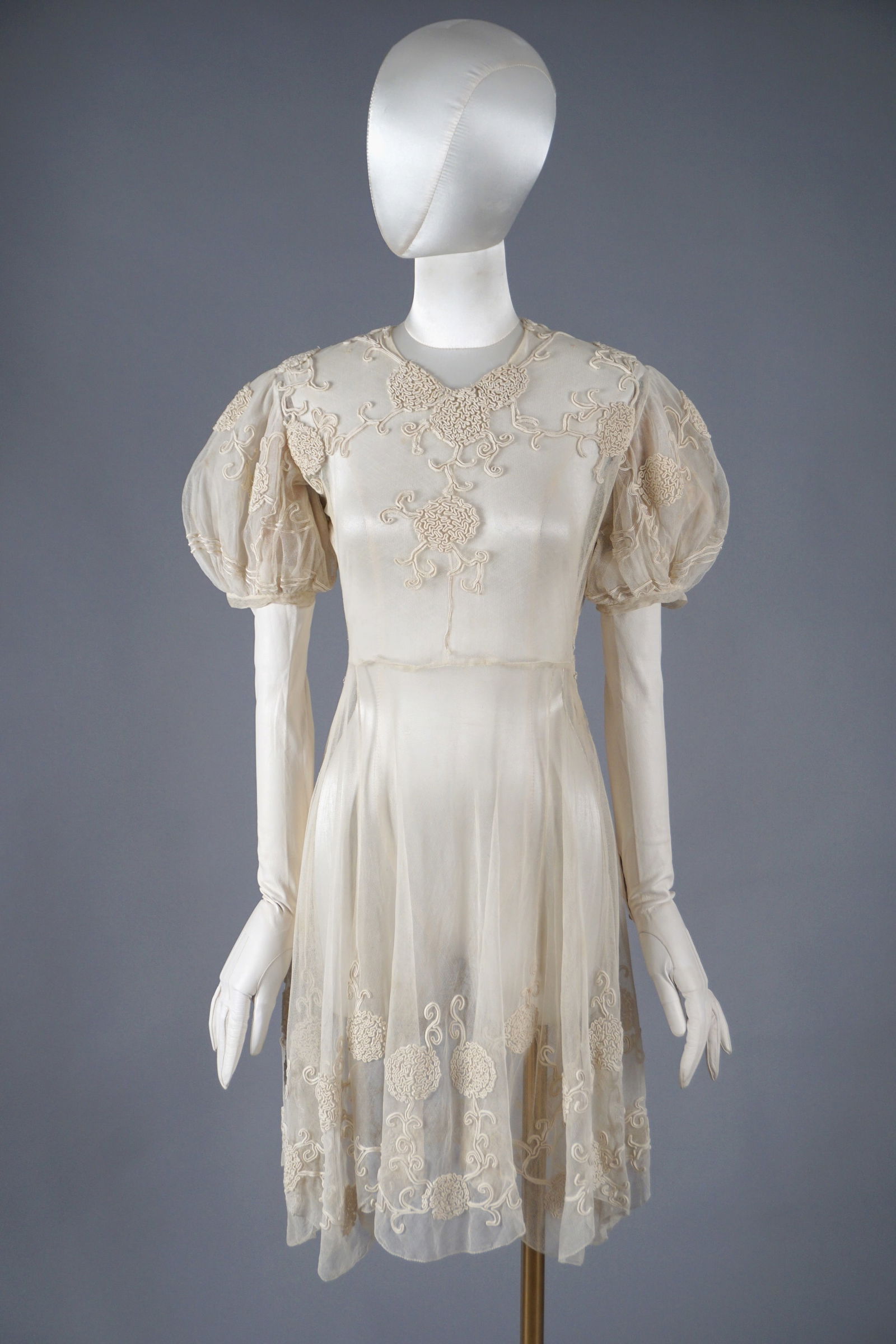 Vintage 1930s Ivory Net Lace Dress with Corded Floral Embroidery: An ethereal 1930s ivory net dress featuring intricate corded floral embroidery throughout. The dress has softly gathered puff sleeves, a delicate neckline, and a short flowing skirt with embroidered m