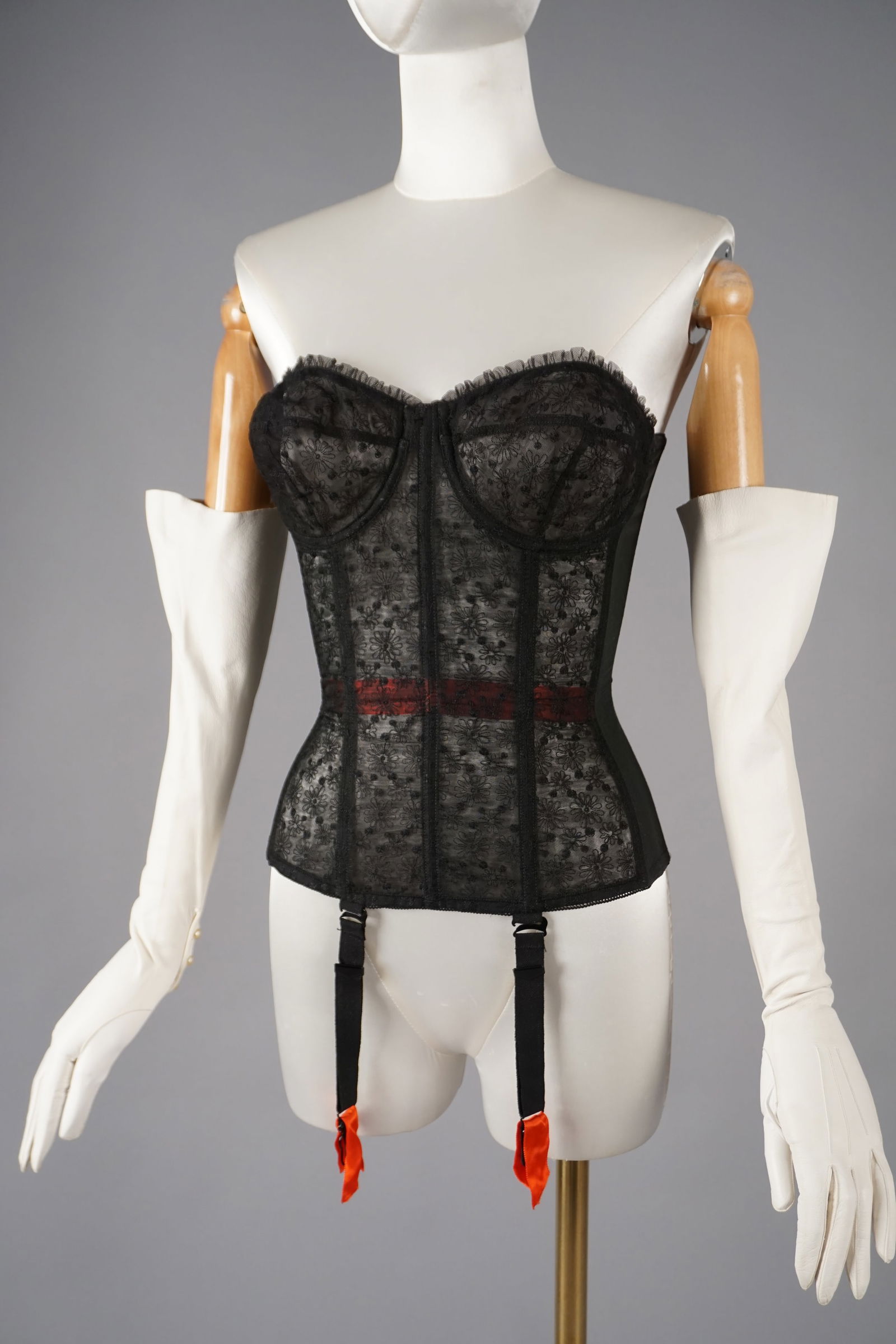 Vintage Black Lace Bustier Corset with Red Garters: A vintage Warner original bustier corset, crafted in sheer black lace with a delicate daisy motif and boned interior. The design features a flattering sweetheart neckline, a deep V front seam, and bol