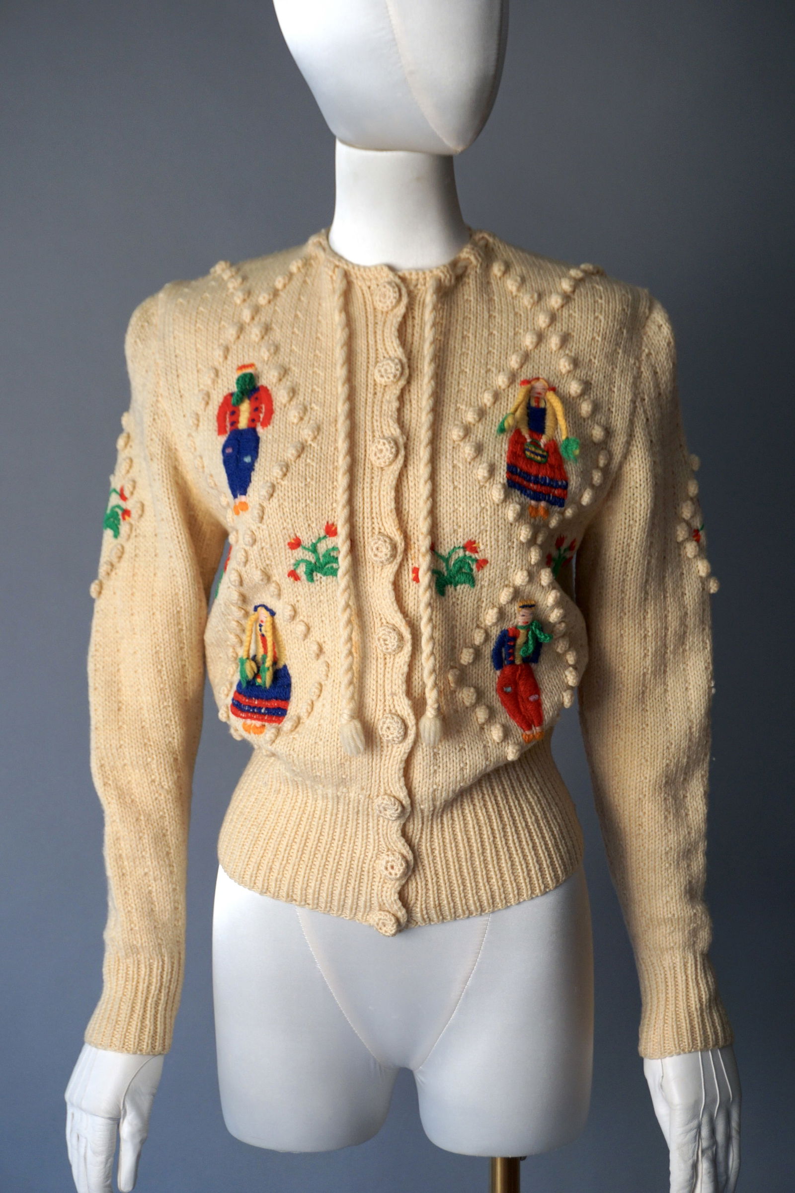 Vintage 1940s Hand Knitted Figural Cardigan Made in England: Charming 1940s hand knitted cardigan is crafted from 100% pure wool in a soft ivory tone. The front is adorned with whimsical figural motifs of men and women in bright folkloric dress, each hand embro