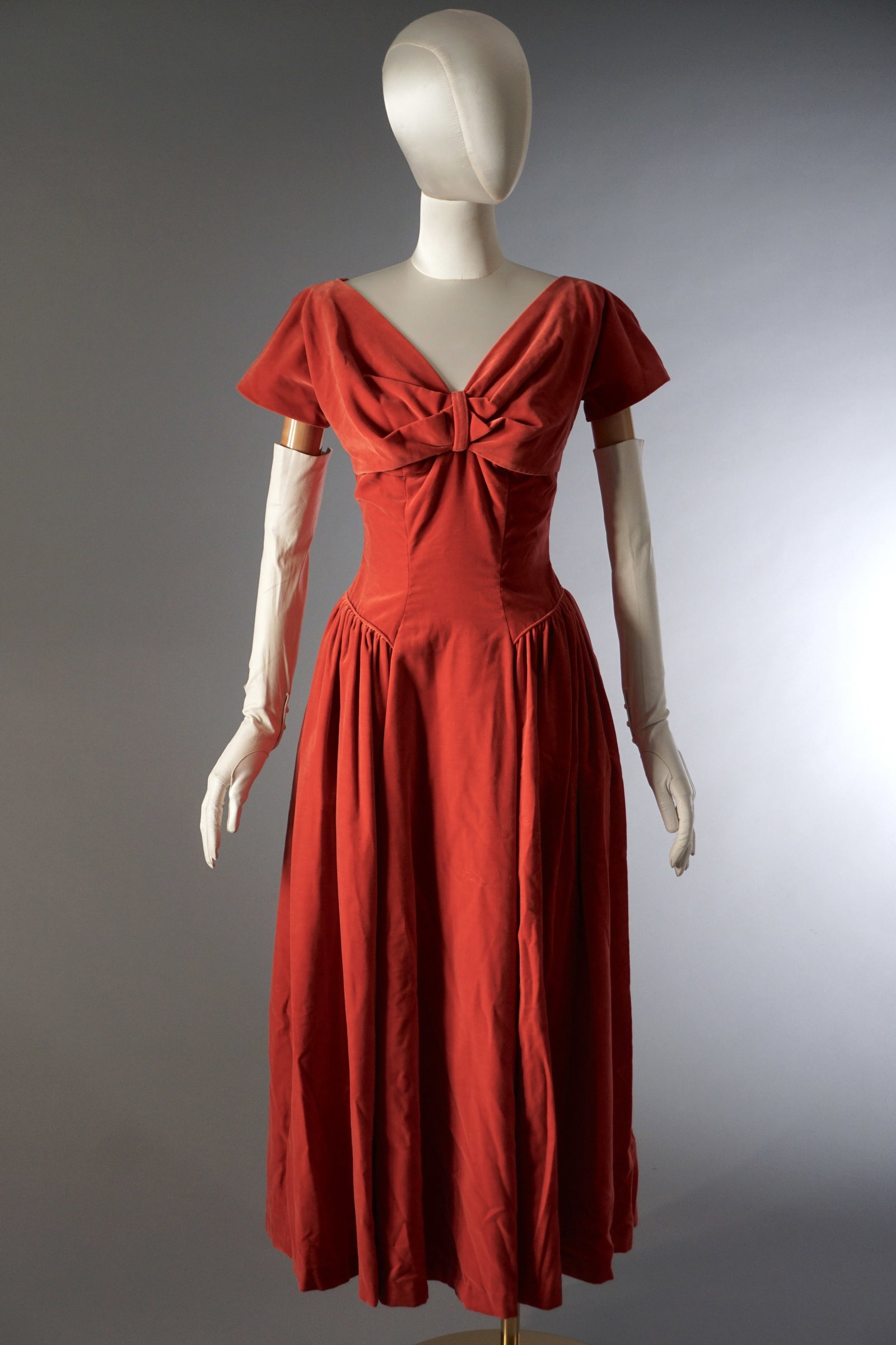 Vintage 1950s Pink Velvet Dress with Bow Front Bodice: A mid century velvet dress in a warm pink tone, shaped with a fitted bodice and a soft, gathered skirt. The neckline with a structured bow detail across the front. Short sleeves and a defined waistlin