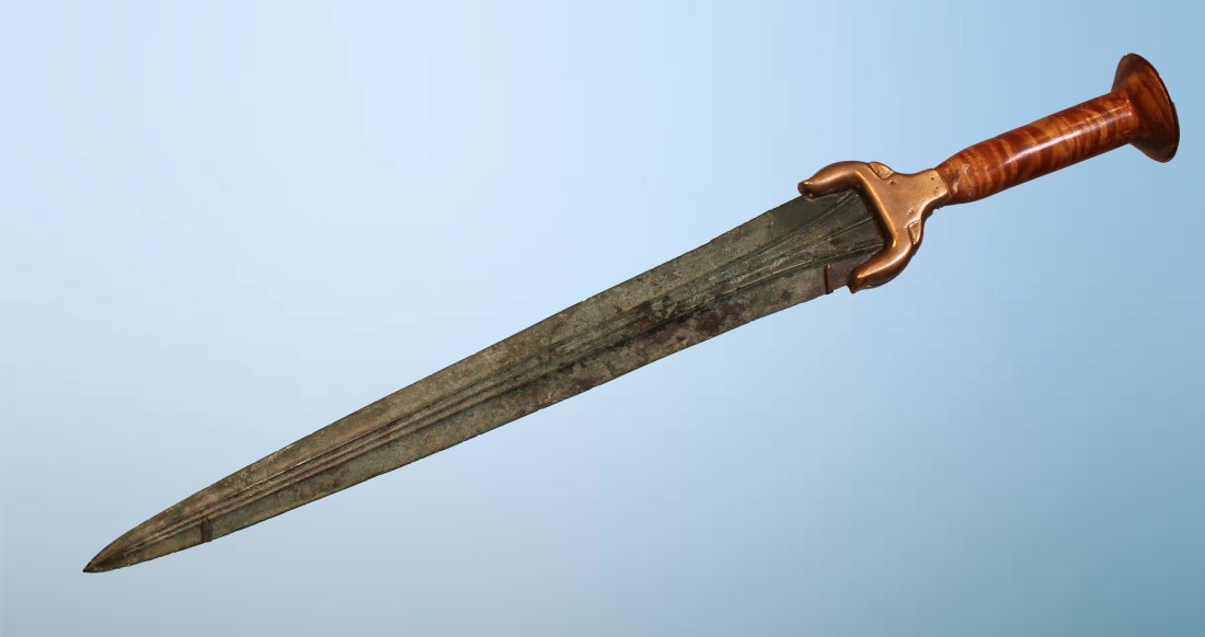LURISTAN BRONZE SWORD: An ancient Luristan bronze sword with a long tapering doubled edged blade with a prominent mid-rib. The blade is set in modern reconstructed hilt of burlwood with a bronze bull head form cross guard.