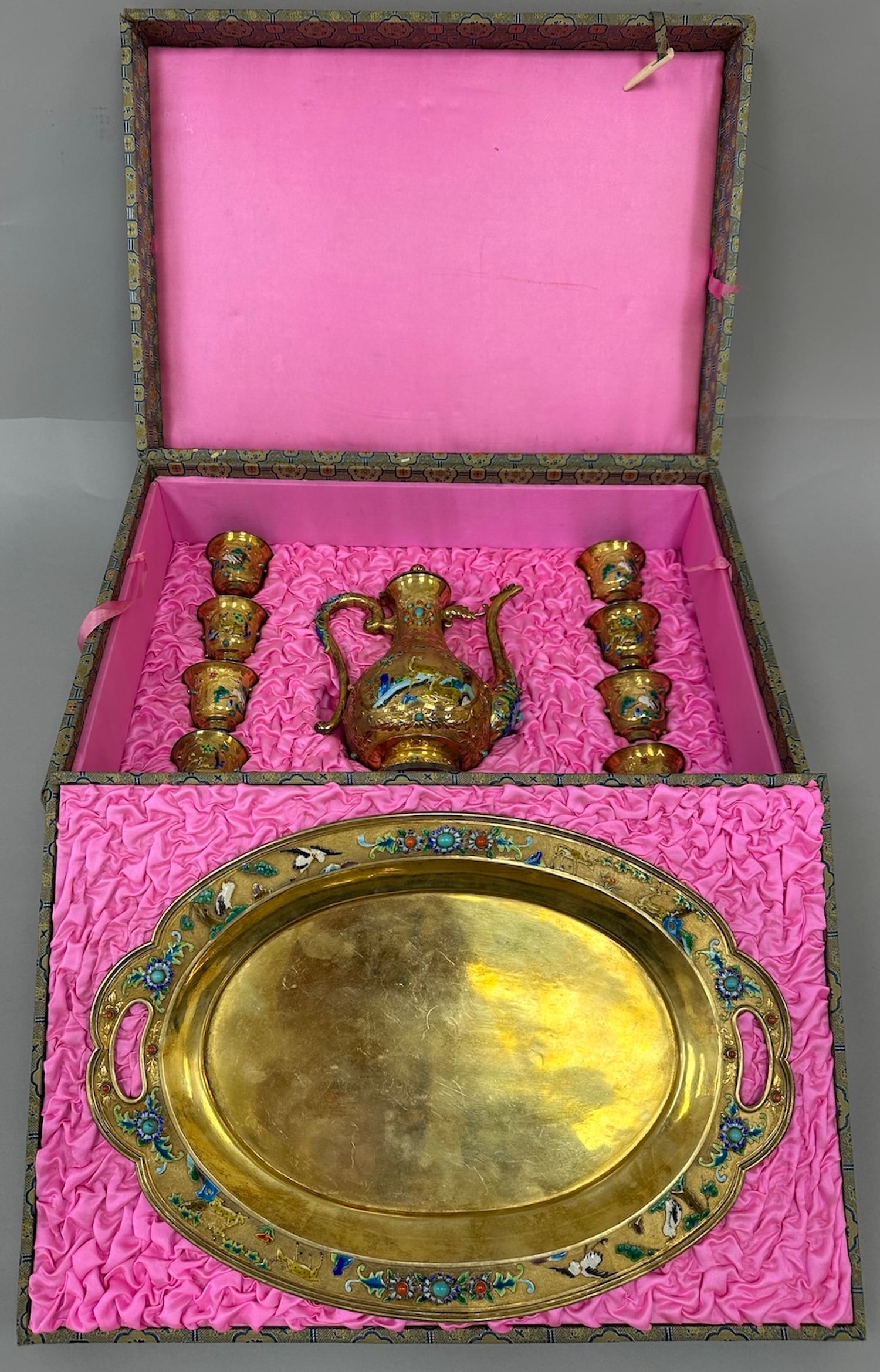 Chinese Bronzed Gilt Enameled Wine Set: Chinese Bronzed Gilt Enameled Wine Set nestled in a pink silk-lined box. The set includes: A wine ewer with a handle and spout, decorated with indicate enamel scenes. 6 small goblets, a large oval tra