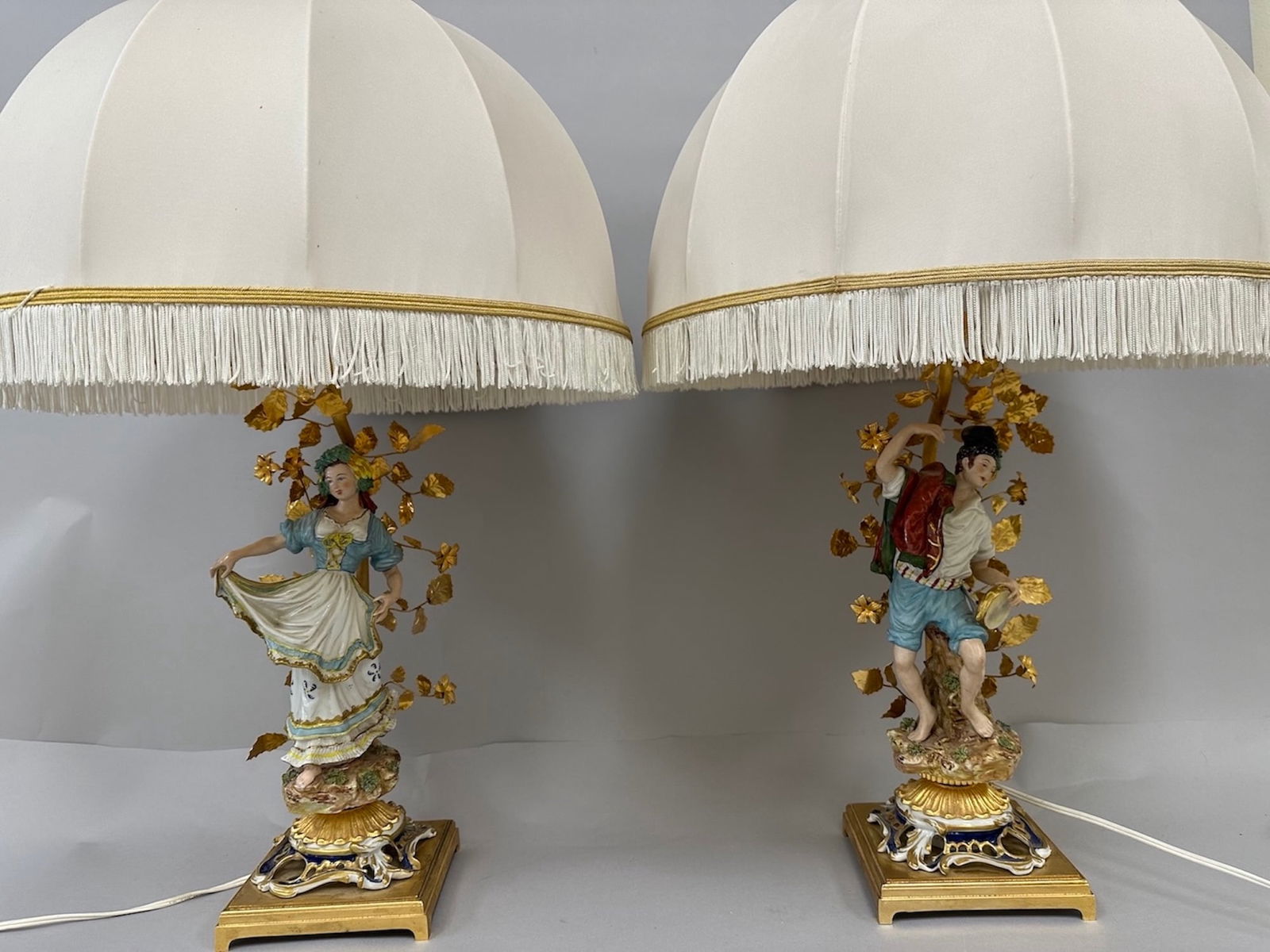 Pair of 2 - 18th Century Style Porcelain Lamps.: A pair of porcelain lamps featuring figurines in 18th - century style attire. The lamps depict a man and a women each standing on an ornate base and surrounded by gilded vines. Each lamp has a white p