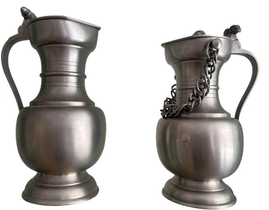 Set of two jugs: 1: Silvered Metal water or milk jug with solid decorated handle and feet. Dimensions: 21,3x14 cm. Dimenisions: circa 23x12 cm. 3: Water or milk jug with decorated lid and original chain from