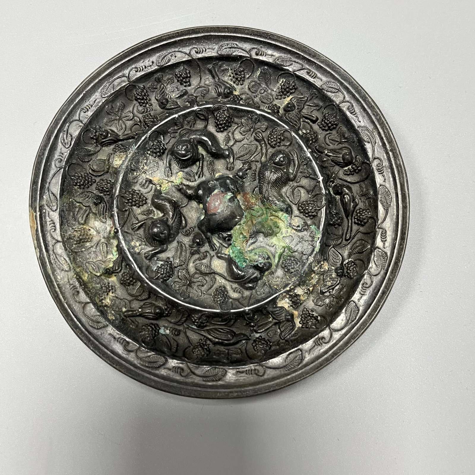 Tang Dynasty Bronze Mirror with Auspicious Beast and Grape Design: This Tang Dynasty bronze mirror with auspicious beast and grape motif measures 11.5cm in diameter and 0.9cm in thickness. It is perfectly round, and its surface, oxidized over a millennium, has develo