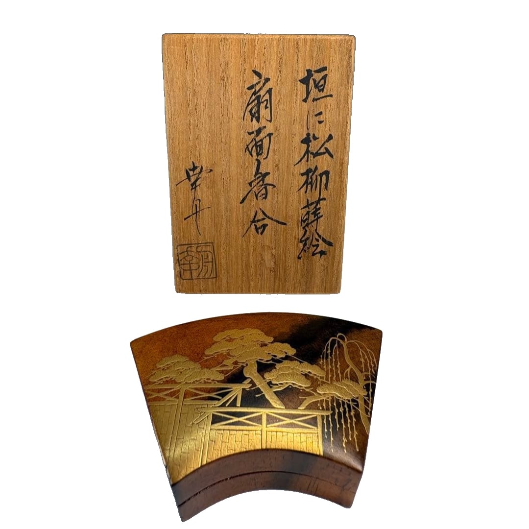Tea Ceremony Utensils: A fan-shaped incense box made of KOshU black persimmon wood, adorned with: This is a fan-shaped incense box, the body of which is adorned with a fence pattern, utilizing the maki-e technique (gold lacquer) to depict motifs of pine and willow trees. This is an item that has