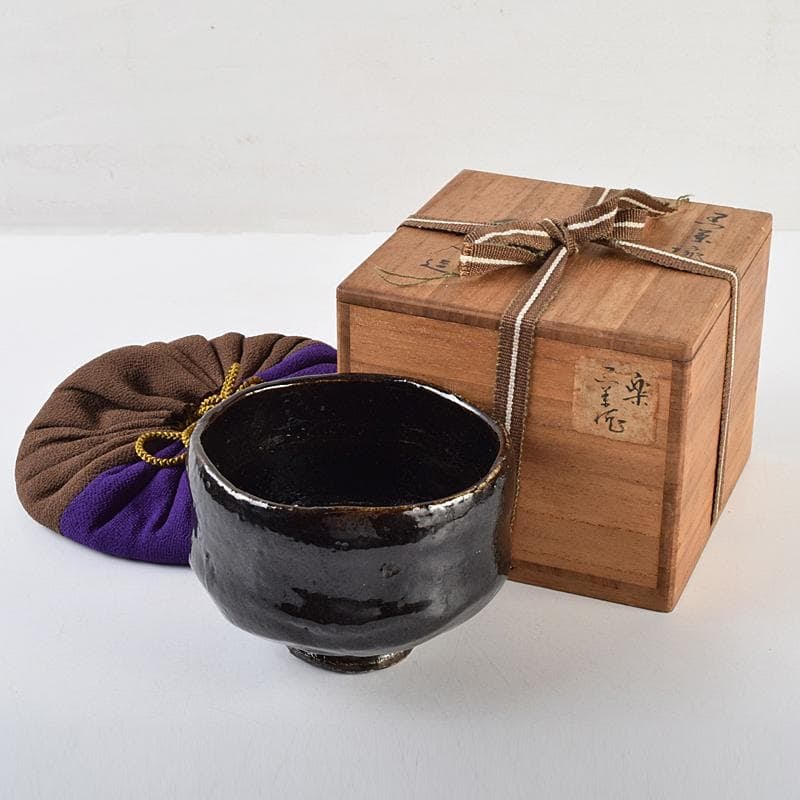 Tea Ceremony Utensils: A black pottery tea bowl crafted by Kichizaemon Hiroshi, accompanied by a: Tea Ceremony Utensil: A black pottery tea bowl crafted by Kizaemon Hiroshi, accompanied by a protective cloth cover and original box. Dimensions: Approximately 8.5 cm in height and 12 cm in width.
