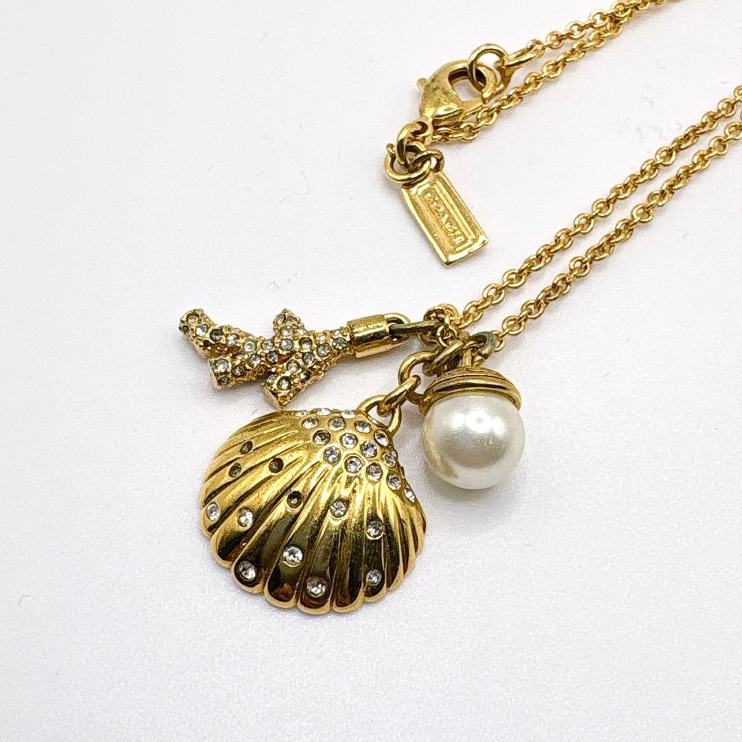 Second-hand - Coach seashell pearl pendant gold necklace (shell style): Brand: Coach Product Description: A charm necklace that combines Coach's signature playfulness with elegant charm. The shell pattern is adorned with sparkling crystals, and a pearl-like pendant hangs 