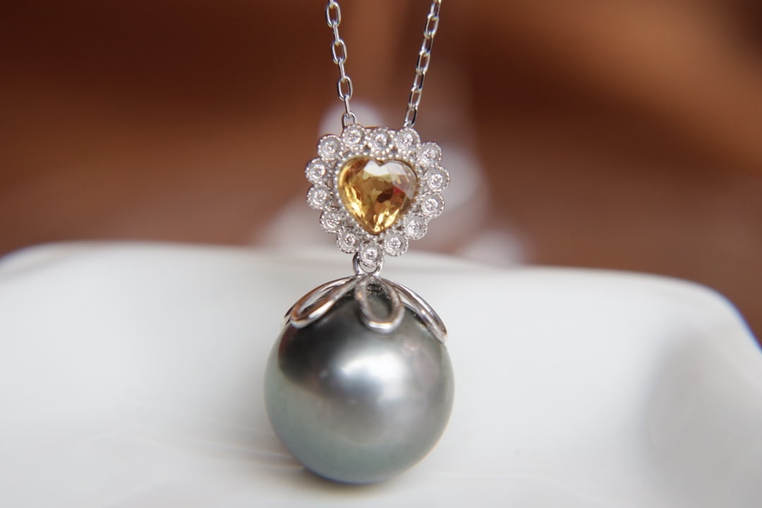 18K Gold Tahitian Pearl Pendant (chain not included): This 18K gold Tahitian pearl pendant features a perfectly round, high luster pearl at its core, emitting a charming allure. The slightly flawed pearl is paired with natural yellow sapphires and blue