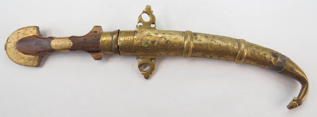 A MOROCCAN KOUMIYYA DAGGER: Mounted in engraved brass, with flared pommel and curved blade, in its matching scabbard. Embellished overall. 19th century. Worn. Overall length 42 cm. Condition