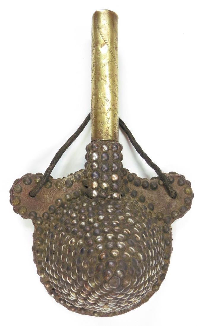 A MOROCCAN POWDER FLASK: The wooden body of characteristic domed form with projecting suspension tabs, covered entirely in leather and heavily embellished with brass studs. The spout covered in embellished brass. First half o