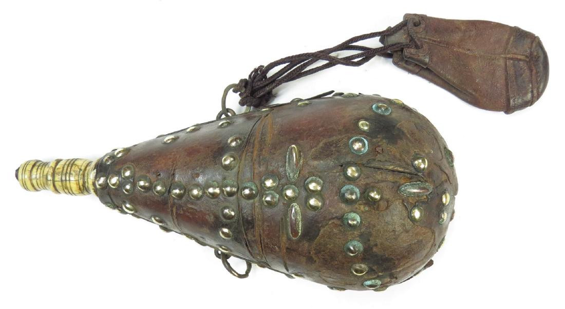 A MOROCCAN POWDER FLASK: The wooden body of characteristic lozenge form, covered entirely in leather and embellished with brass studs and turned spout. With bullet bag intact First half of the 19th century. Light wear. Overal