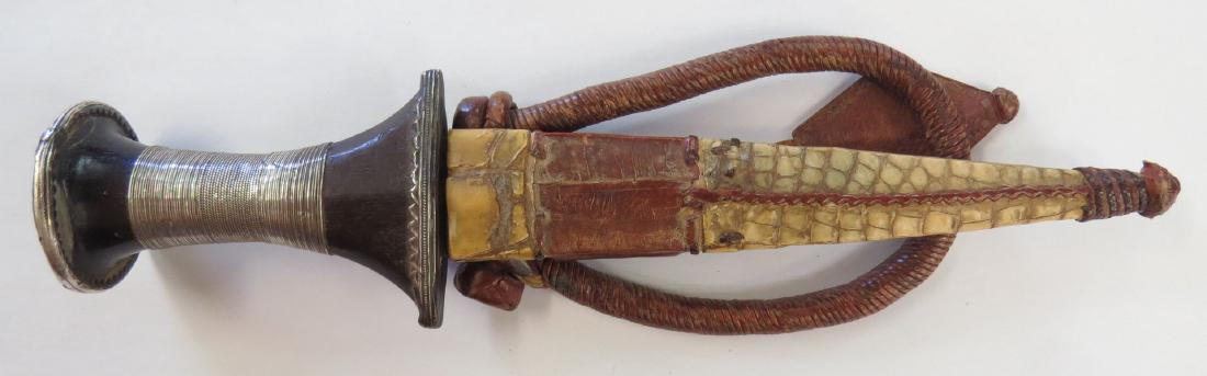 A FINE SUDANESE ARM DAGGER: The carved ebony hilt with silver pommel and ferrule, the grip wrapped with finely braided silver wire. The straight, double-edged blade retaining some original edge, the leather scabbard with white c