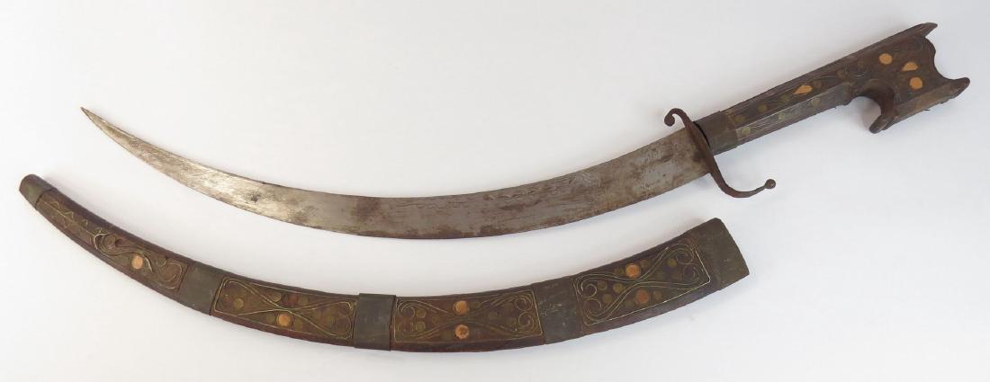 AN ALGERIAN NIMCHA DAGGER: The wooden mounts carved overall with brass bands. With characteristic scrolled and hooked pommel and recurved guard. The curved blade with engraved motifs. 20th century. Overall length 62.4 cm. Condi
