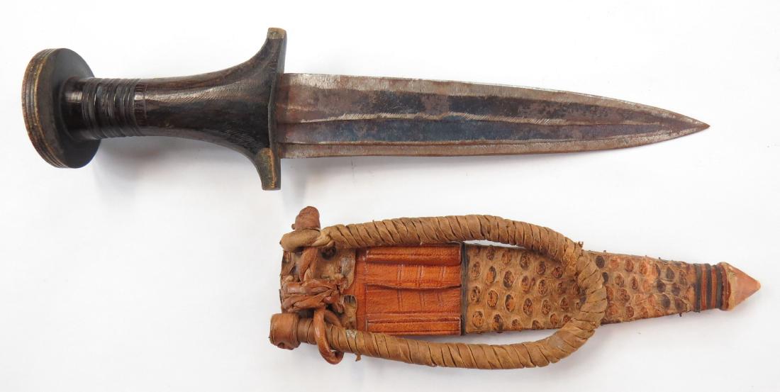 A SUDANESE ARM DAGGER: The characteristic grip carved of ebony, with straight, double-edged, fullered blade. In its tooled leather sheath with lizard tip and wrist strap intact. 19th-20th century. Worn. Overall length 31cm.
