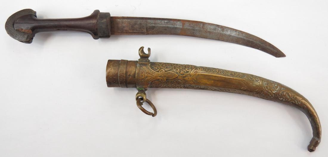 A MOROCCAN KOUMIYYA DAGGER: Mounted in engraved brass, with flared pommel and curved blade, in its matching scabbard. 19th century. Well worn, lug damaged, tip lacking. Overall length 41.5 cm. Condition IV