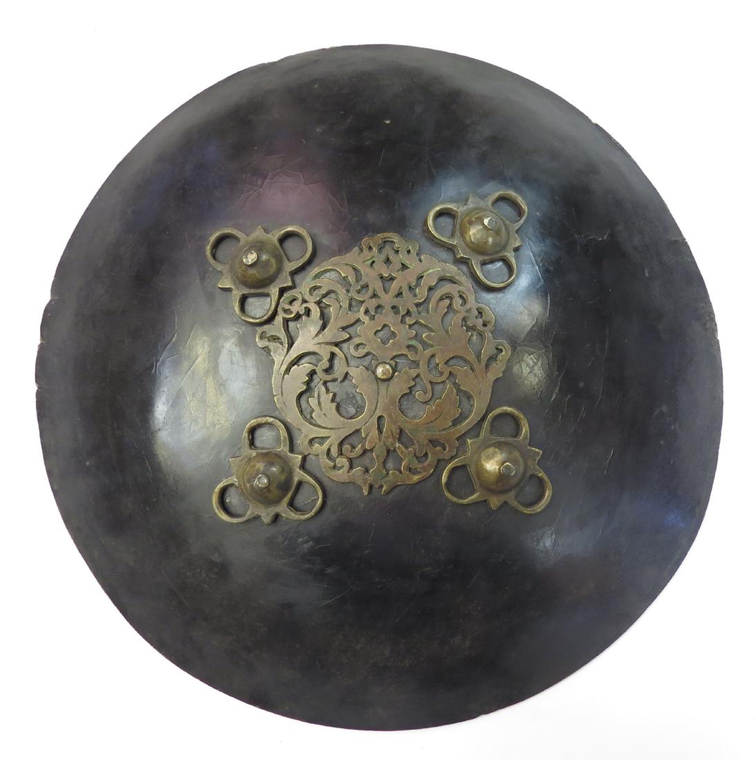 AN INDIAN DHAL SHIELD: An elaborate example formed of lustrous elephant hide, deeply domed. With applied pierced and scrolled central brass boss, and four pierced brass bosses. 18th-19th century. Worn. Diameter 40cm. Condit