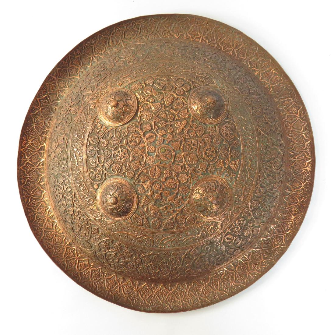 A RARE UZBEK SHIELD: Wrought entirely of copper and superbly embossed and chased with Central Asian flowering vine motifs and inscriptions, with four raised bossed and rolled edge. Grips intact to reverse. 19th century. L