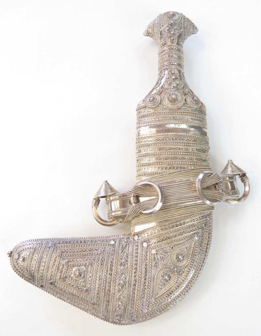 AN OMANI KHANJAR DAGGER: Of typical form, the mounts wrought entirely of filigreed and embossed silver. The curved, double-edged blade with central rib, wooden scabbard with heavy silver mounts en suite with the hilt. 20th ce