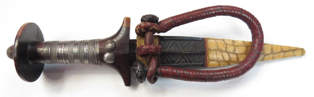 A FINE SUDANESE ARM DAGGER: The carved ebony hilt embellished with finely braided silver wire and profiled studs. The straight, double-edged blade retaining some original edge, the leather scabbard with white crocodile mounts, a