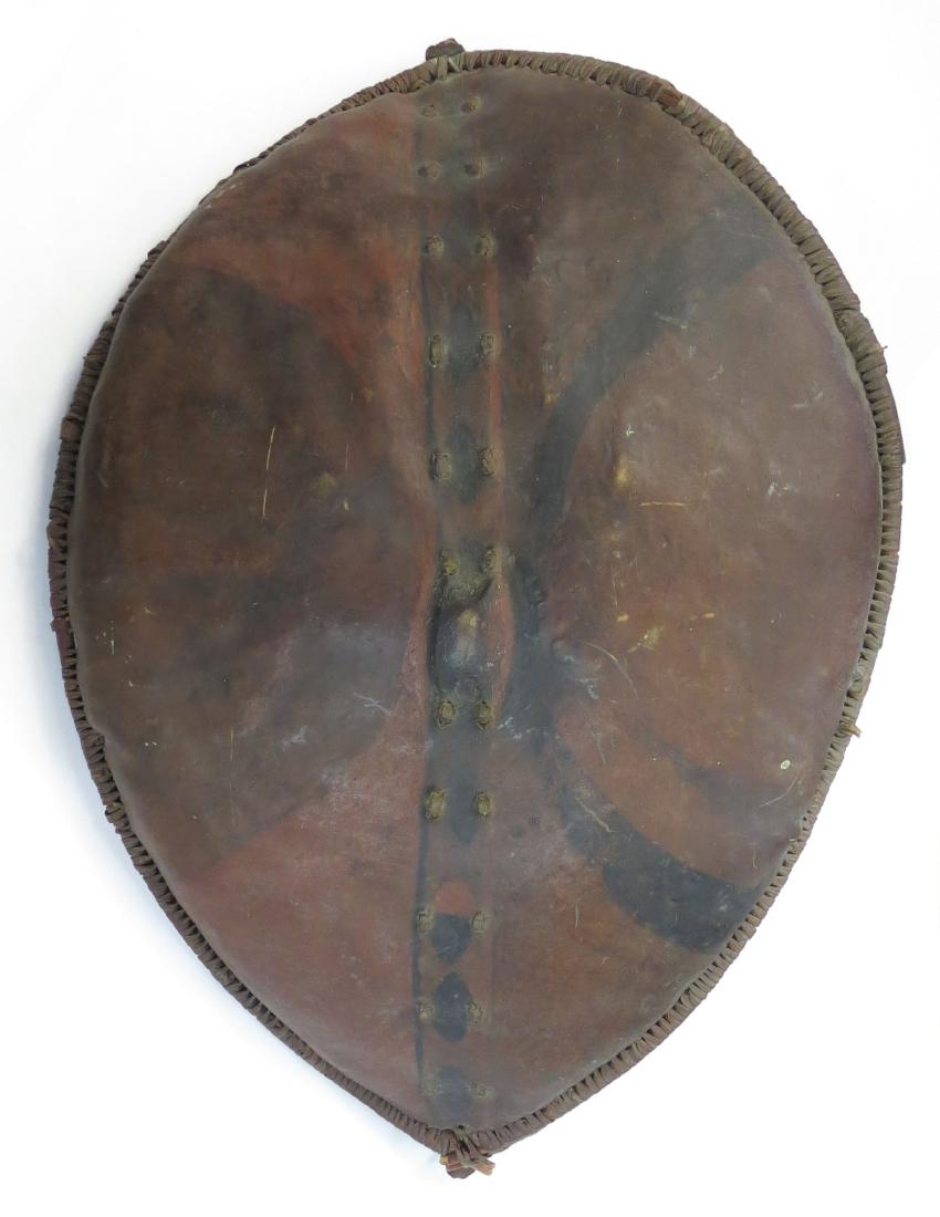 A RARE MAASAI SHIELD: A large example of characteristic form, made of hide laced to a central wooden post with integral grip, the edges bound to stripped branches and hammered-iron rods with tightly-coiled hide. The obvers