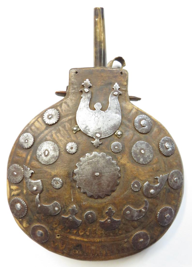 A RARE CENTRAL ASIAN POWDER FLASK: Of circular form, carved of wood and covered with tooled hide, set with numerous profiled and engraved steel ornaments. With spout, cutoff and suspension loops intact. 18th-19th century. Minor losses.