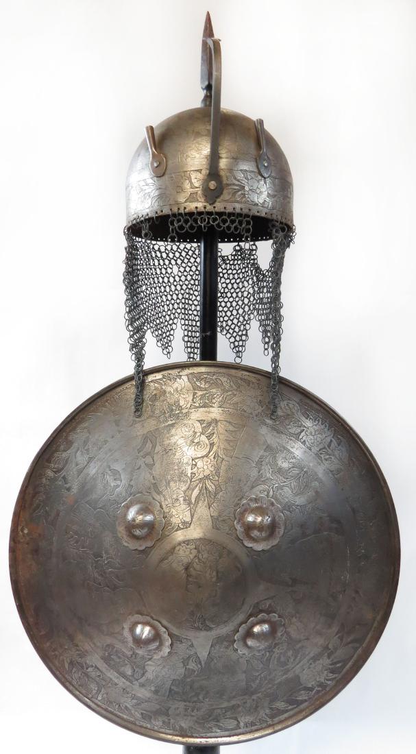A PERSIAN ARMOR SET: Comprised of an embellished helmet and shield, etched with scenes from Persian mythology. The helmet of characteristic hemispherical form with elongated top spike, nasal bar and plume tubes, embellish