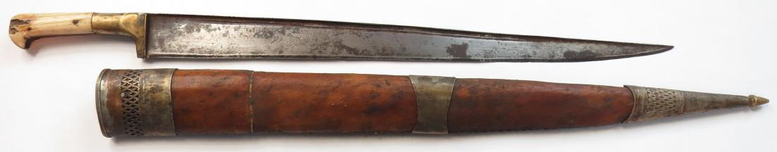 AN AFGHAN KHYBER KNIFE: A large example, with elephant grips and faceted bolster, the broad steel blade wrought with a T-back. In its leather-covered wooden scabbard with brass mounts. 19th century. Worn, with minor losses. 