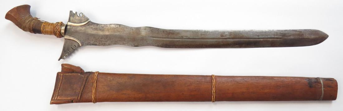 A MORO SWORD KRIS: Tausug People, with characteristic flared pommel, rattan binding largely lacking. The waved, fullered, handforged blade with silver baca-bacas. In its fragmented wooden scabbard. Late 19th -early 20th