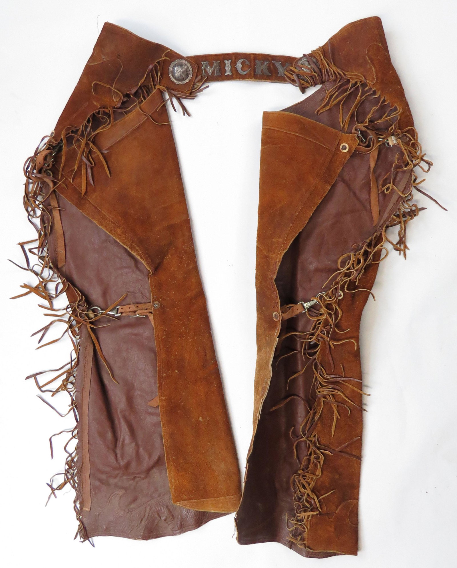 A PAIR OF AMERICAN WESTERN CHAPS: Roughout, elaborately fringed and set with finely-engraved silver conchos and profiled and engraved letters MICKY. Mid-20th century. Light wear. Overall length 103cm. Condition II