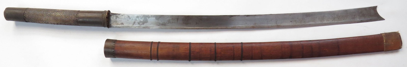A BURMESE DHA: The hilt with brass mounts and rayskin-wrapped grip, the well-wrought, hollowground blade with cusped tip. In its hardwood scabbard with brass mounts. 19th-20th century. Minor wear. Overall length 75.